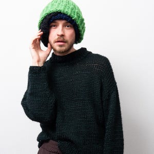 May include: A dark green knitted jumper with a high neckline and long sleeves. The model is wearing a green, blue, and black knitted beanie and brown trousers. The jumper has a chunky knit texture.