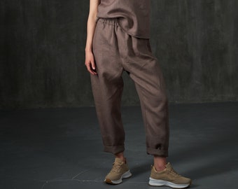 May include: A woman is wearing a brown linen jumpsuit with a relaxed fit. The jumpsuit has a scoop neck and thin straps. She is wearing beige sneakers.
