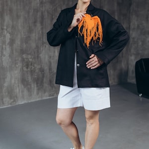Puede incluir: Una mujer con un blazer negro, pantalones cortos blancos y zapatillas blancas. Lleva una bufanda naranja brillante en la mano.