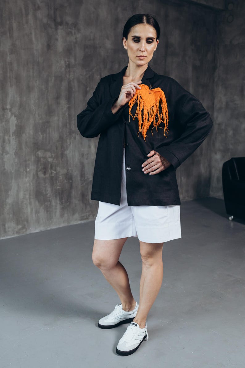 Puede incluir: Una mujer con un blazer negro, pantalones cortos blancos y zapatillas blancas. Lleva una bufanda naranja brillante en la mano.