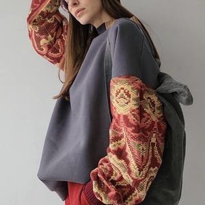 Può includere: Una felpa grigia con un berretto bordeaux e pantaloni rossi. Le maniche della felpa hanno un design a fantasia colorata.