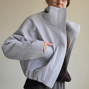 Może przedstawiać: Jas pikowana w jasnoniebieskim kolorze z wysokim kołnierzem i wzorem w kratkę. Kurtka zapinana na zamek błyskawiczny, z kieszenią po lewej stronie.