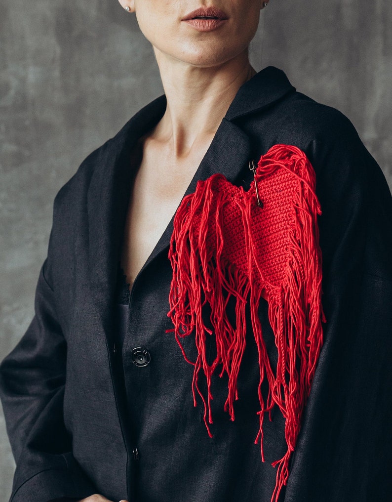 Puede incluir: Una chaqueta negra con un broche de coraz&oacute;n rojo tejido a crochet con detalles de flecos.