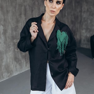 Puede incluir: Una mujer con un blazer de lino negro con un broche verde en forma de coraz&oacute;n y pantalones cortos de lino blanco.