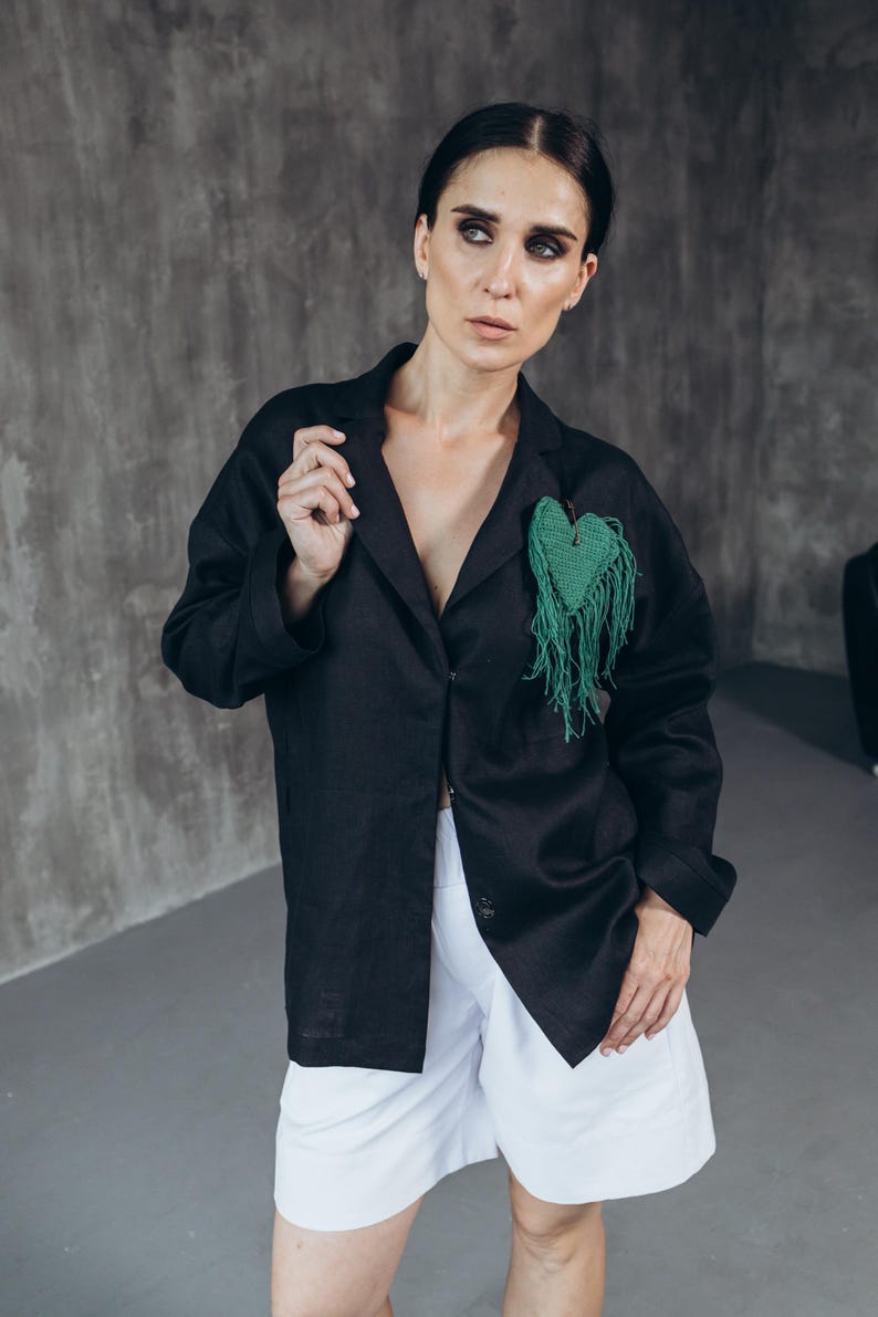 Puede incluir: Una mujer con un blazer de lino negro con un broche verde en forma de coraz&oacute;n y pantalones cortos de lino blanco.