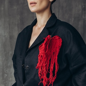 Puede incluir: Una chaqueta negra con un broche de coraz&oacute;n rojo con flecos. El broche est&aacute; hecho de hilo rojo y tiene un borde deshilachado.