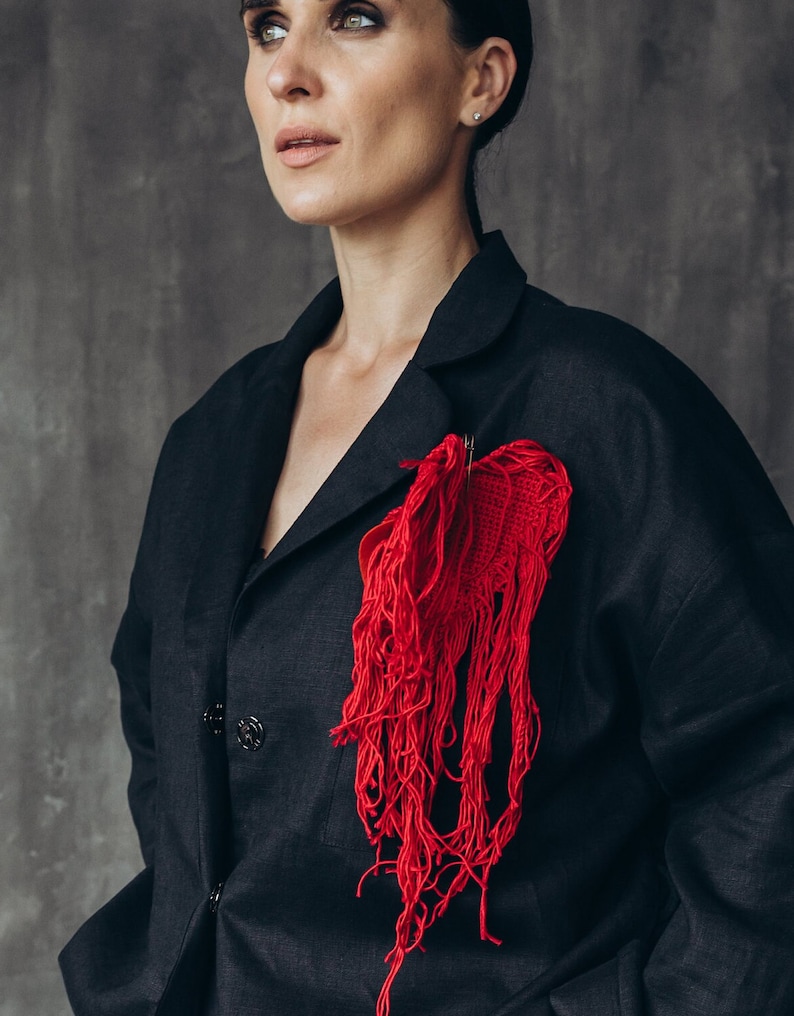 Puede incluir: Una chaqueta negra con un broche de coraz&oacute;n rojo con flecos. El broche est&aacute; hecho de hilo rojo y tiene un borde deshilachado.