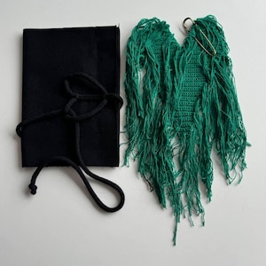 Puede incluir: Una pieza de crochet de forma de coraz&oacute;n de color verde con flecos, unida a un imperdible. Tambi&eacute;n se muestra una bolsa de tela negra con cord&oacute;n.