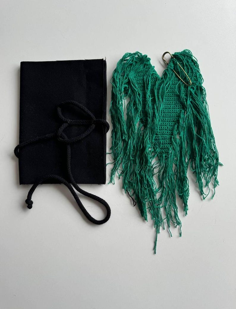 Puede incluir: Una pieza de crochet de forma de coraz&oacute;n de color verde con flecos, unida a un imperdible. Tambi&eacute;n se muestra una bolsa de tela negra con cord&oacute;n.