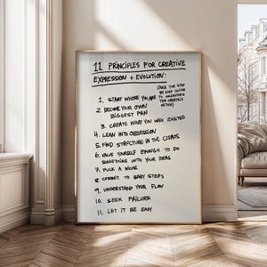 以下が含まれることがあります： 「11の創造的表現+進化の原則」と手書きで書かれたテキストがフレームに入っています。テキストは黒インクで白い背景に書かれています。フレームは金色です。プリントは、窓とソファのある壁に立てかけられています。