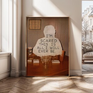 May include: A framed print with the text "I'M SCARED THIS IS ALL I WILL EVER BE." The artwork shows a person's silhouette with the text. The print is in a gold frame, with a wooden table and other objects in the foreground.
