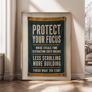 Op de afbeelding: Een rechthoekige donkerblauwe stoffen wandhanger met beige randen en metalen ringen in de hoeken. De tekst luidt: PROTECT YOUR FOCUS, NOISE STEALS TIME, DISTRACTION COSTS DREAMS, LESS SCROLLING MORE BUILDING, FINISH WHAT YOU START in beige.