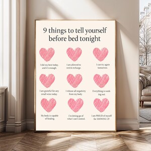 May include: A framed print with the text "9 things to tell yourself before bed tonight." The print features nine pink heart illustrations, each paired with a positive affirmation. The frame is a warm brown color, and the background is a light beige.