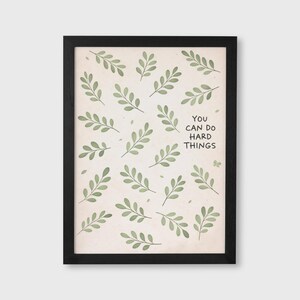 May include: A framed art print featuring a beige background with scattered green leaf illustrations. The black frame surrounds the artwork, which includes the text "YOU CAN DO HARD THINGS" in black font.