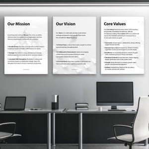 May include: A modern office setting with three framed posters displaying company values: "Our Mission," "Our Vision," and "Core Values." The posters are white with black text. Desktops and office chairs are also visible.