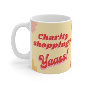 May include: White ceramic mug with a yellow background and abstract pink and orange shapes. The text "charity shopping? Yaass!" is written in red with a retro font.