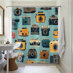 May include: A teal shower curtain featuring a repeating pattern of vintage cameras in various colors, including yellow, blue, and brown. The cameras have a retro design, with lenses and dials. The curtain is hanging in a bathroom with a white sink, towels, and a rug.