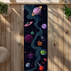 Peut inclure: Un tapis de yoga noir avec un imprimé galaxie représentant une nébuleuse bleue, des étoiles et des planètes dans des tons de rose, violet, vert et jaune.