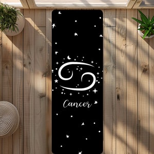May include: Black yoga mat with a white Cancer zodiac sign and stars. The sign is surrounded by a circle of stars. The text "Cancer" is below the sign.