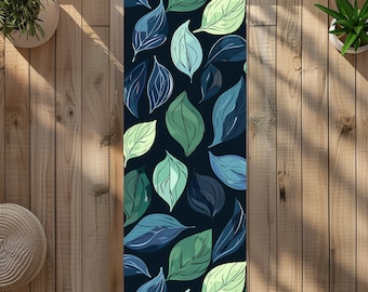 Könnte beinhalten: Rechteckige Yogamatte mit einem Muster aus stilisierten Blättern in Blau- und Grüntönen auf dunklem Hintergrund. Die Blätter haben weiße Umrisse. Die Matte liegt auf einer Holzoberfläche.