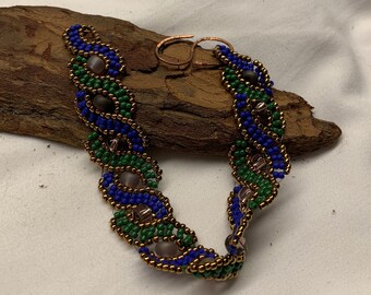 May include: A beaded bracelet with a copper clasp. The bracelet features a woven design of blue, green, and brown beads. The bracelet is resting on a piece of wood against a white background.