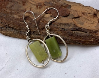 Può includere: Un paio di orecchini fatti a mano con pietre rettangolari verde oliva incastonate in cornici ovali argentate. Gli orecchini hanno ganci in filo d'argento. Il design è semplice ed elegante, adatto all'uso quotidiano.