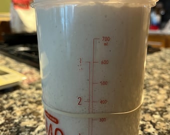 May include: A clear plastic container with a red lid and a rubber band around the middle. The container has markings for measuring in milliliters, up to 700 ml. The container is filled with a white substance, likely sourdough starter.