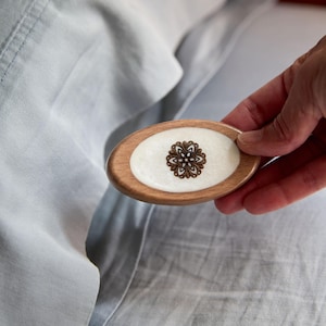 May include: An oval wooden hair barrette with a white center and a brown floral design. The barrette is held against a light blue fabric background.
