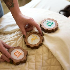 May include: Four wooden chakra symbols with different colored Sanskrit symbols and designs. The symbols are on a white and beige quilted surface.