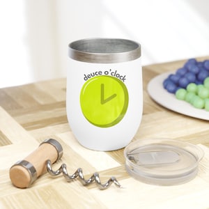 May include: White stainless steel wine tumbler with a green clock face and the text "deuce o'clock". The tumbler is sitting on a wooden surface with a corkscrew and a clear lid.