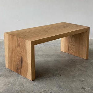 Peut inclure: Une table basse rectangulaire fabriquée en bois naturel de couleur claire avec un motif de grain visible. La table présente un design en cascade où la surface supérieure se prolonge en deux panneaux latéraux verticaux continus.