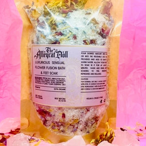 Op de afbeelding: Een doorzichtige plastic zak gevuld met een bad- en voetbad. Het bad is een mengsel van Epsom-zout, Himalaya-zout, zout van de Dode Zee, baking soda, calendula, lavendel, rozenblaadjes, rozen-geranium, patchouli en ylang-ylang etherische oliën. De zak is gelabeld met "The Alleycat Doll Luxurious Sensual Flower Fusion Bath & Feet Soak". Het label vermeldt ook "100% biologisch" en "Vegan & Cruelty Free".