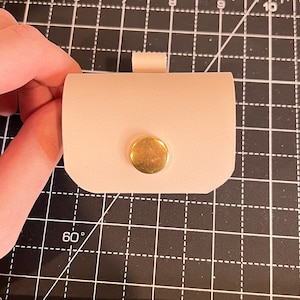 Puede incluir: Una pequeña bolsa de cuero rosa claro con un cierre de botón de metal dorado. La bolsa está sobre una estera de corte negra.