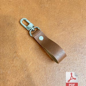 May include: A brown leather keyring with a silver-coloured clasp and rivet. The keyring is made from a single strip of leather and is designed to hold keys or other small items. The background is a textured brown surface.