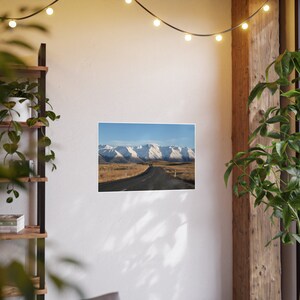 May include: A winding road leads through a valley with snow-capped mountains in the distance. The road is paved and surrounded by brown grass.