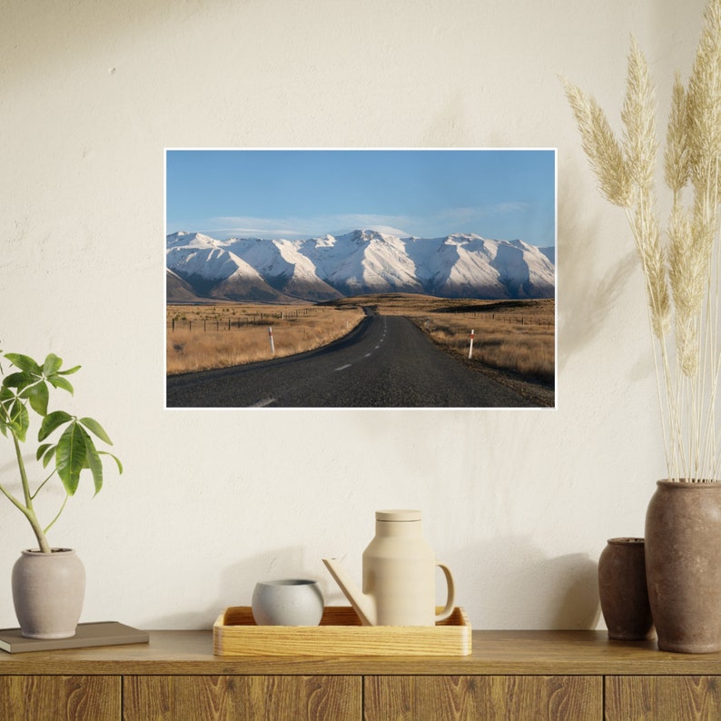 May include: A winding road leads towards snow-capped mountains under a clear blue sky. The road is paved and surrounded by brown grass and a few trees.