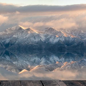 May include: A scenic view of snow-capped mountains with a wooden plank floor in the foreground. The mountains are covered in white snow and the sky is a soft pink and orange.