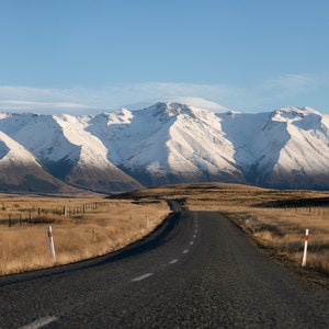 May include: A long, paved road stretches towards a snow-capped mountain range in the distance. The road is surrounded by dry, brown grass and a fence.