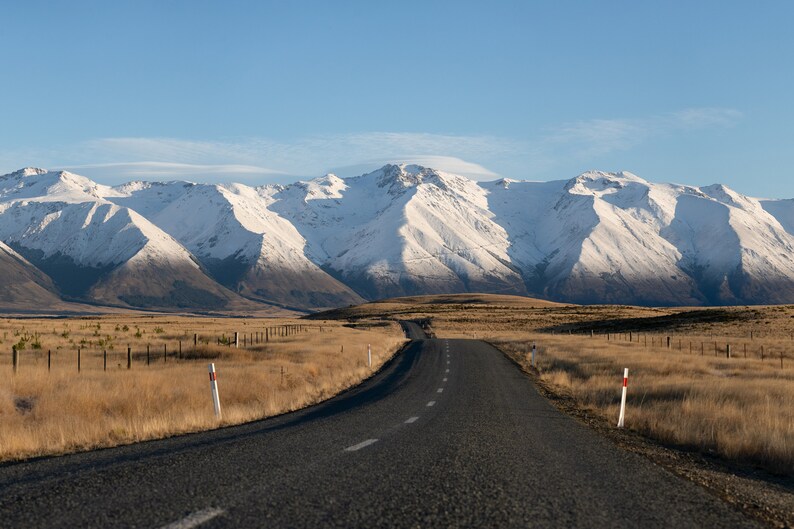 May include: A long, paved road stretches towards a snow-capped mountain range in the distance. The road is surrounded by dry, brown grass and a fence.