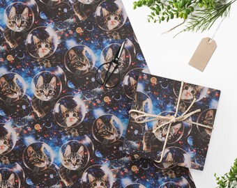 Könnte beinhalten: Geschenkpapier mit einem sich wiederholenden Muster von Katzen, die Astronautenhelme tragen, vor einem Galaxie-Hintergrund. Die Katzen sind in verschiedenen Posen und haben unterschiedliche Gesichtsausdrücke. Das Papier ist blau, schwarz und weiß.