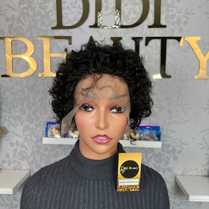 Puede incluir: Peluca rizada negra con frente de encaje en un maniquí. El maniquí lleva un jersey de cuello alto gris. El fondo presenta las letras doradas "DIDI BEAUTY" sobre una pared con estampado floral.