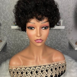 Puede incluir: Una peluca corta y rizada negra con raya lateral. La peluca está peinada con un corte pixie y se lleva en una cabeza de maniquí. El maniquí lleva una blusa negra de hombros descubiertos con ribete de encaje dorado.