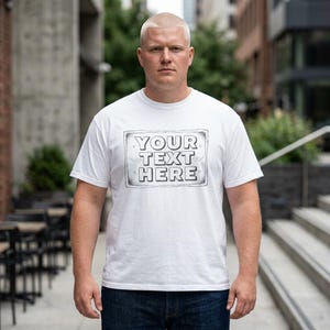 Puede incluir: Una camiseta blanca de manga corta con un gráfico rectangular en el pecho que contiene el texto YOUR TEXT HERE en contorno negro. El diseño incluye pequeños iconos de alas decorativas a los lados del texto dentro del borde.