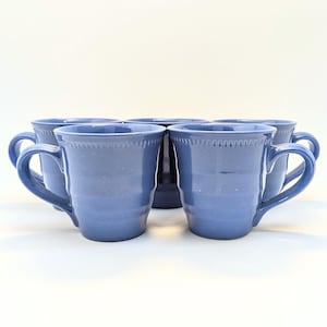 May include: A set of six blue ceramic mugs with curved handles. The mugs have a speckled texture and a decorative beaded rim. The mugs are arranged in a semi-circle on a white surface.