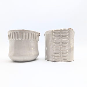 May include: Two white ceramic planters with different embossed designs. The planter on the left has a textured rim and the planter on the right has a repeating oval pattern.