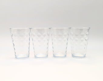 May include: Four clear drinking glasses with a textured, dotted pattern. The glasses are cylindrical with a slightly flared top and sit on a white surface. The glasses are empty and appear to be made of glass.