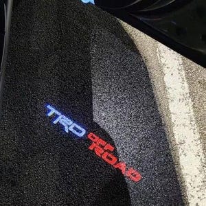 Puede incluir: Alfombrilla de coche negra texturizada con el texto iluminado "TRD" en azul y "OFF ROAD" en rojo. La alfombrilla está en ángulo, mostrando una parte de la puerta del coche y el borde de la carretera.