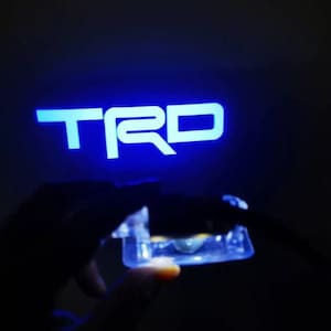 Puede incluir: Logotipo "TRD" azul brillante sobre un fondo oscuro. Un objeto rectangular pequeño y transparente se sostiene frente al logotipo. El objeto parece estar iluminado.
