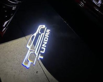 Puede incluir: Una brillante proyección de luz LED blanca en el suelo que muestra el contorno de una camioneta y la palabra TUNDRA en letras mayúsculas. La proyección es nítida y clara sobre la superficie oscura, mostrando el diseño del logo.