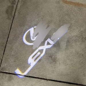 Puede incluir: Una brillante proyección de luz blanca del logotipo de Lexus y la palabra LEXUS mostrada sobre un suelo de hormigón gris. La proyección presenta un diseño nítido y de alto contraste con el emblema circular sobre el texto estilizado de la marca.